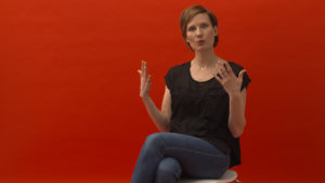 Mutter aus dem Film "Meine Narbe" sitzt vor rotem Hintergrund und spricht über ihre Kaiserschnitterfahrung